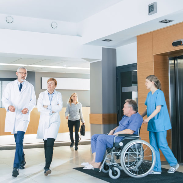 First Floor of the Busy Hospital, Doctors, Nurses and Personnel Busy Working, Assistant Moves Elderly Man in the Wheelchair. New Modern Medical Hospital with Professional Staff.